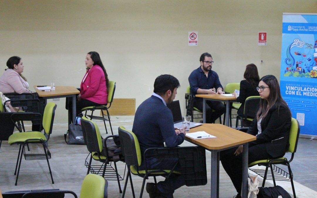 Una fotografía de una jornada de apresto laboral de la Unidad de Vínculos con la Comunidad Egresada y Empleadores. En la foto hay tres mesas, de izquierda a derecha, en donde una persona entrevista a otra en el contexto de entrevistas laborales. al costado derecho de la imagen aparece el pendón celeste de la Unidad.